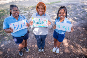 Three students pose holding hand-drawn signs with the words helpful, growth and guidance